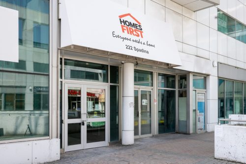 Exterior view of the affordable supportive housing building at 292-222 Spadina Ave. It’s photo of grey building with an entrance sign saying “Homes First, Everyone needs a home”.