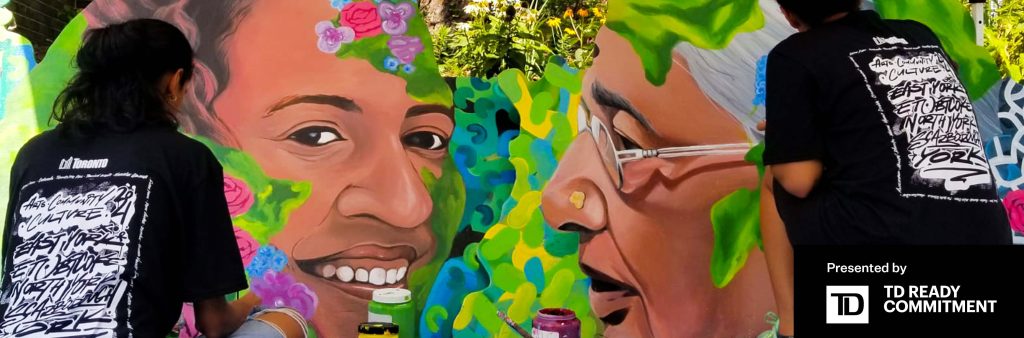 A mural of two oversized portraits surrounded by flowers and greenery is flanked by two artists.