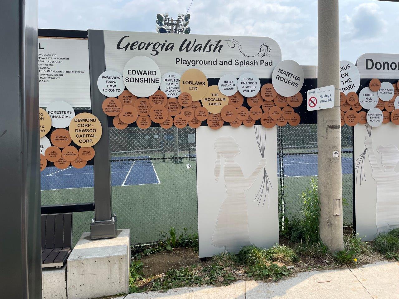 The Donor Wall shown along the exterior fence of the tennis courts showing circular balloon shapes in different sizes and colours with donor names on each balloon. Two silhouettes are shown holding a string connected to the various balloons above.