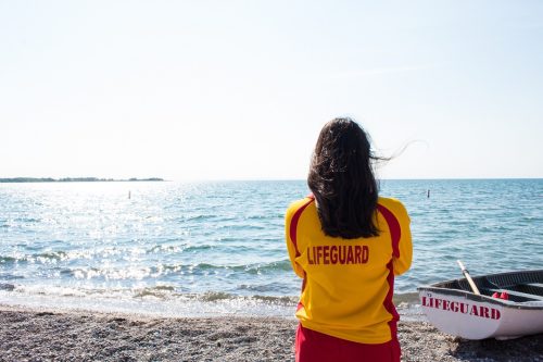 A lifeguard wearing a red-and-yellow uniform surveys the beach. In front of the lifeguard is a white rowboat. 