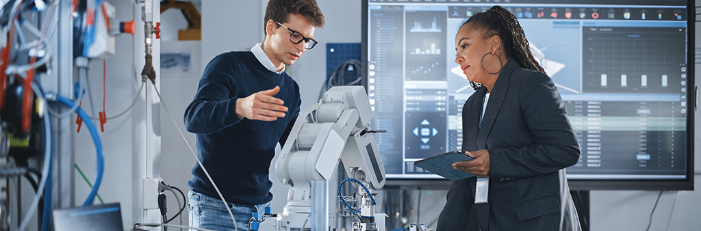 Two people discuss a robotic arm with a large digital display behind them