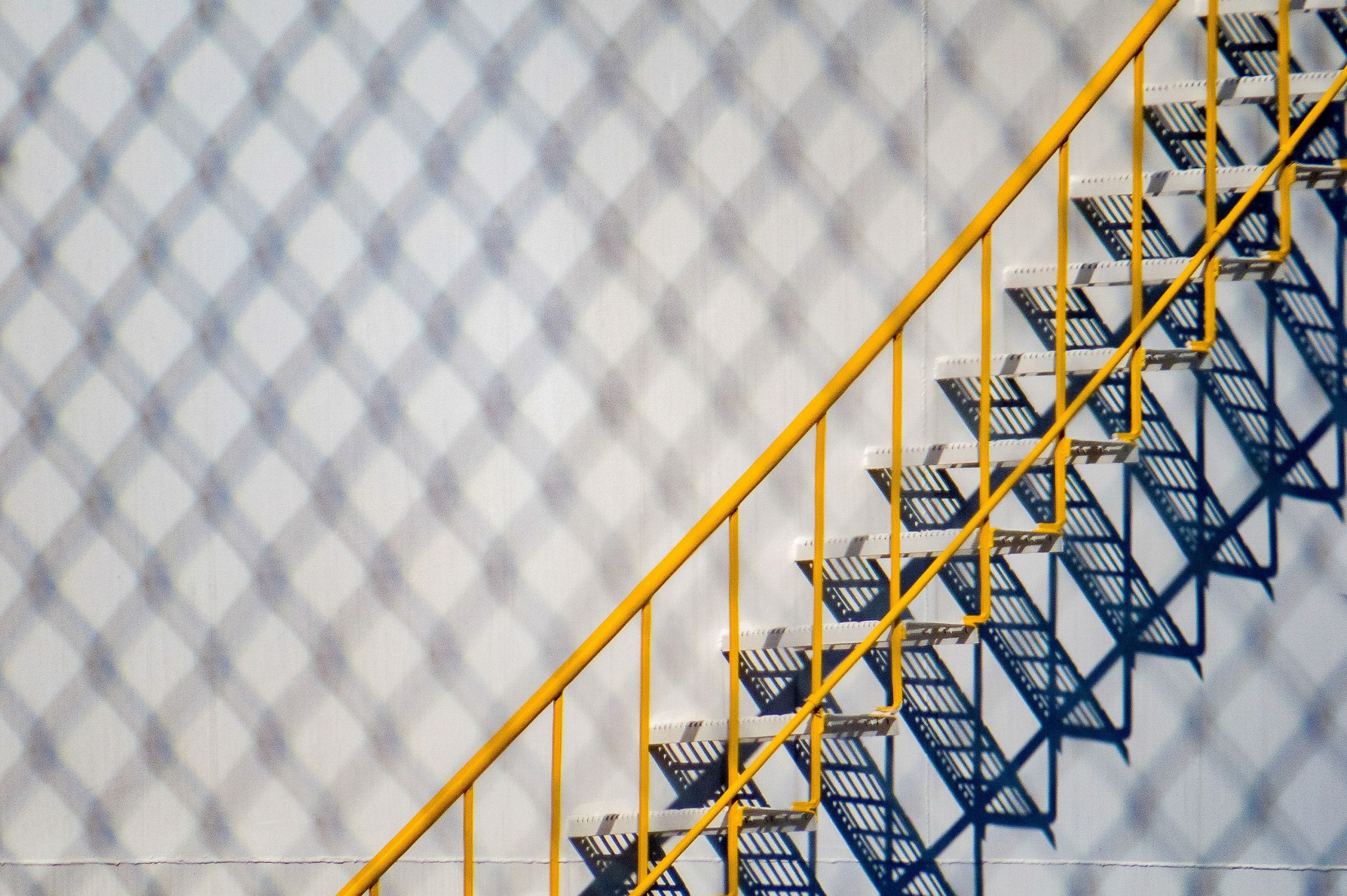 Metal staircase and yellow handrail against a white wall. covered by shadow of chain link fence