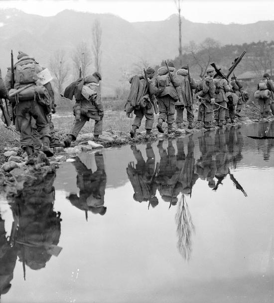 A company of soldiers moving single file across rice paddies