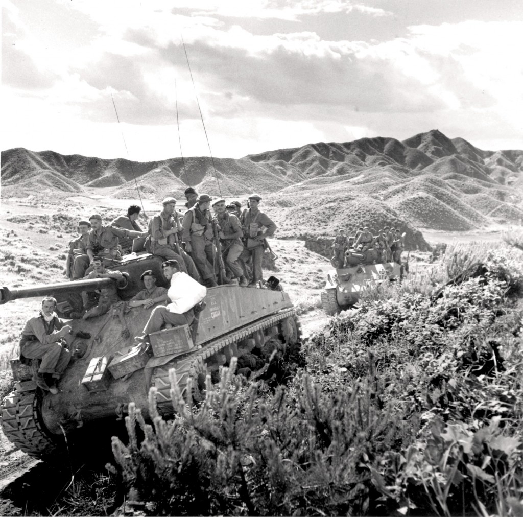 Two tanks travelling down a dirt road, with Canadian soldiers riding on top.