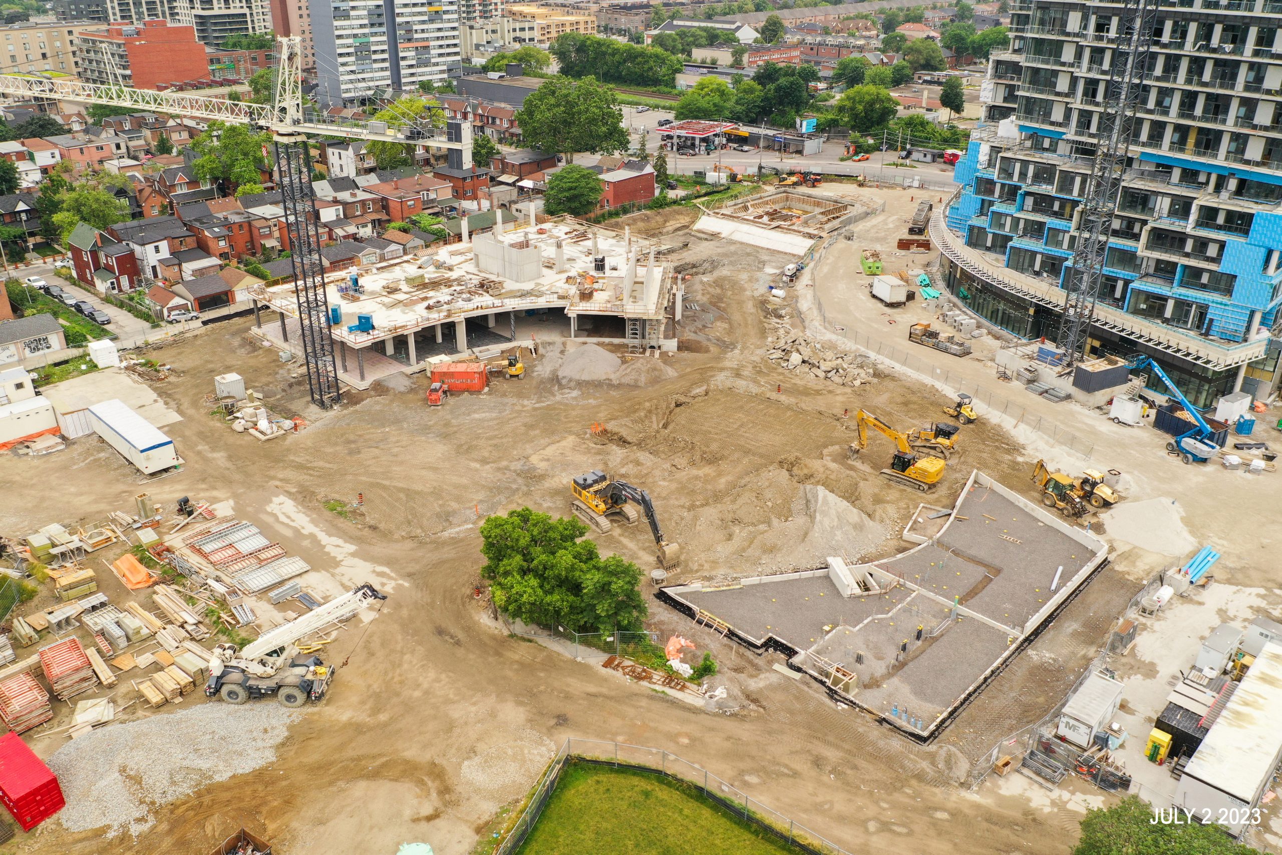An aerial photograph of the construction site at Wallace Emersion Community Centre on July 2, 2023, which focuses on the community centre while under construction and the adjacent Fieldhouse. The community recreation structure is one-storey with vertical beams being installed to the future third floor. The Fieldhouse is at ground-level ready for concrete pouring. 