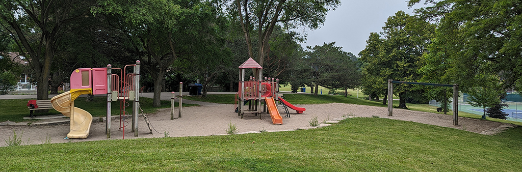 Broadlands Park Playground in the summer with grass in the foreground and two play structures with slides in the background. The playground is surrounded by tall mature trees.