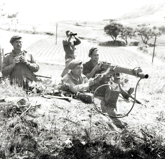 On a hillside, two soldiers operating a machine gun and two behind with binoculars, all looking into the distance.