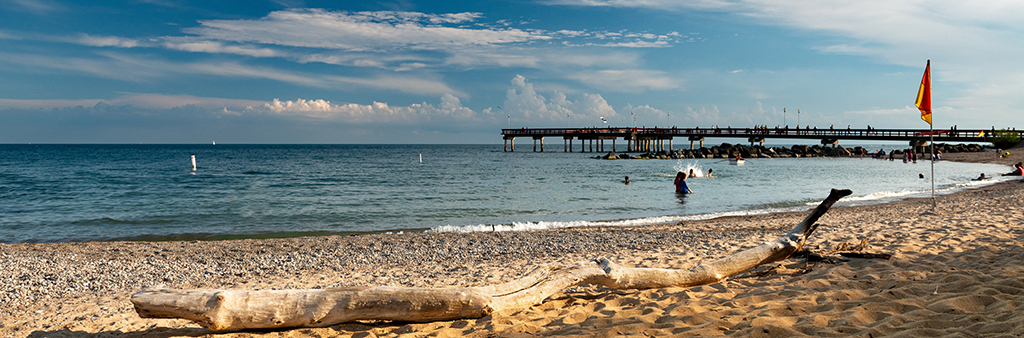 A picture of a beach on a summer day. People are swimming in the water and a lifeguard is watching them from a row boat in the distance. Off into the background is a pier.