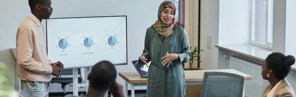 A Black man wearing a peach coloured shirt and a woman wearing a green dress and a head scarf present pie charts to a Black man and woman sitting in a boardroom.