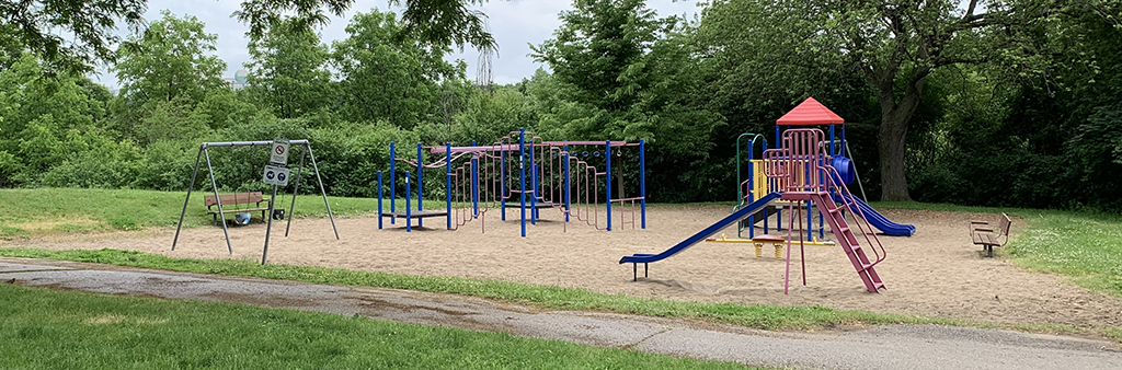 Havenbrook Park Playground which shows various metal play structures in blue and red on top of sand. Mature trees and grass surround the play area, with a narrow concrete path along the perimeter.