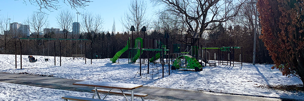 The new Havenbrook playground looking south from the open area to the north of the playground. The playground and park are snow covered with a new picnic table and main walkway in the foreground, and mature vegetation in the background.