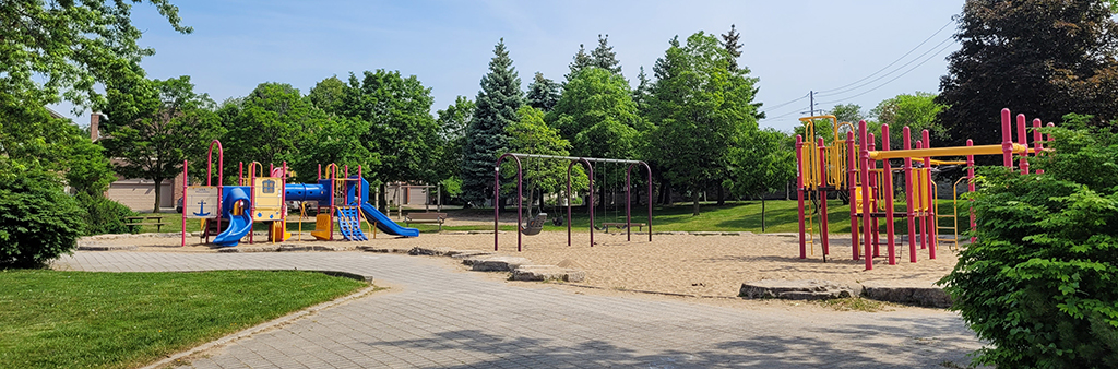 Balmoral Park Playground showing the main pathway to the play area in the foreground, and the playground in the background with various play features, like two slides in blue, a swing set and a structure with climbing features in yellow and red.
