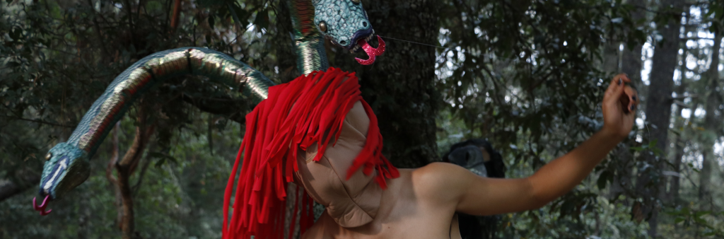 Person wearing headpiece over face and head with two snakes and red hair