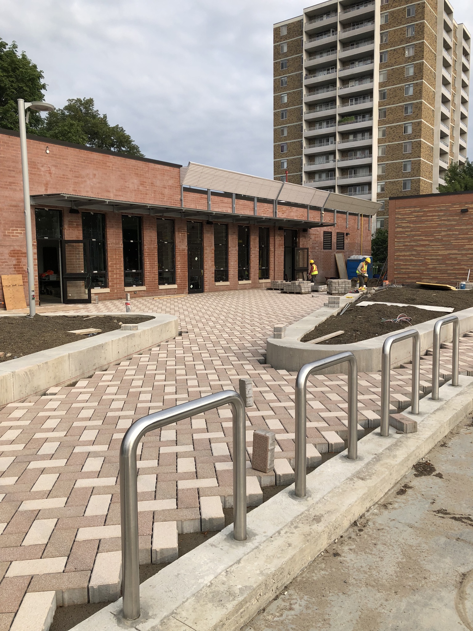 View of public plaza from southeast corner looking northwest, with Clubhouse in background and new paving seen in the foreground. The pavers are two colours, beige and a soft brick colour, and are laid in a herringbone pattern.