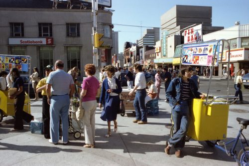 Colour photograph of street corner with crowd of people and ice cream vendor.
