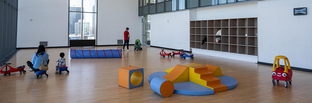 A spacious indoor play area with a high ceiling and large windows, featuring various toys and play equipment. Adults and children are seen interacting with the toys, creating a safe and engaging environment.