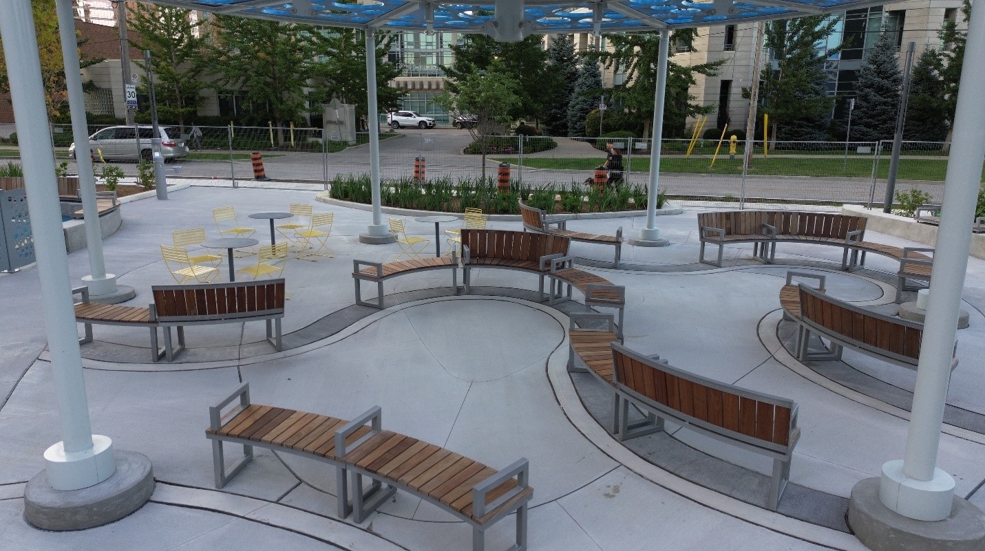 An image of the curvilinear seating area with benches that include arm rests and backs in certain sections. In the background there are small round tables for eating and bright yellow chairs.