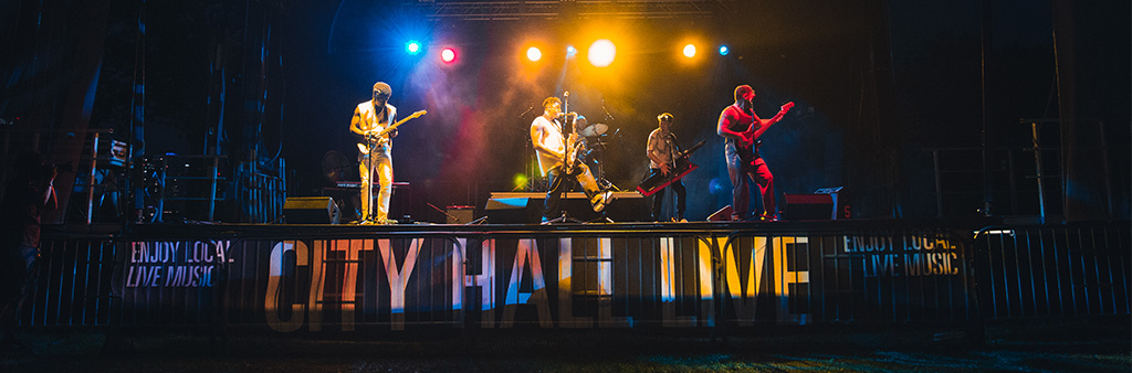 Band playing on outdoor stage at night, banner reads City Hall Live