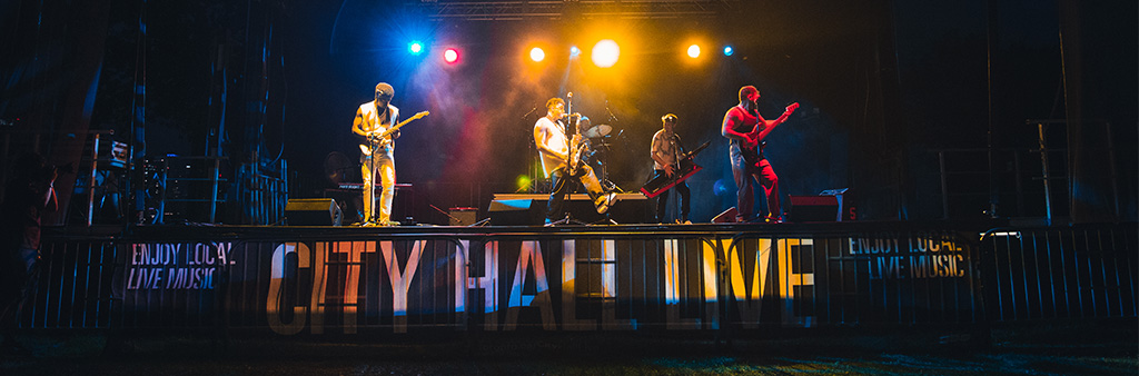 Band playing on outdoor stage at night, banner reads City Hall Live