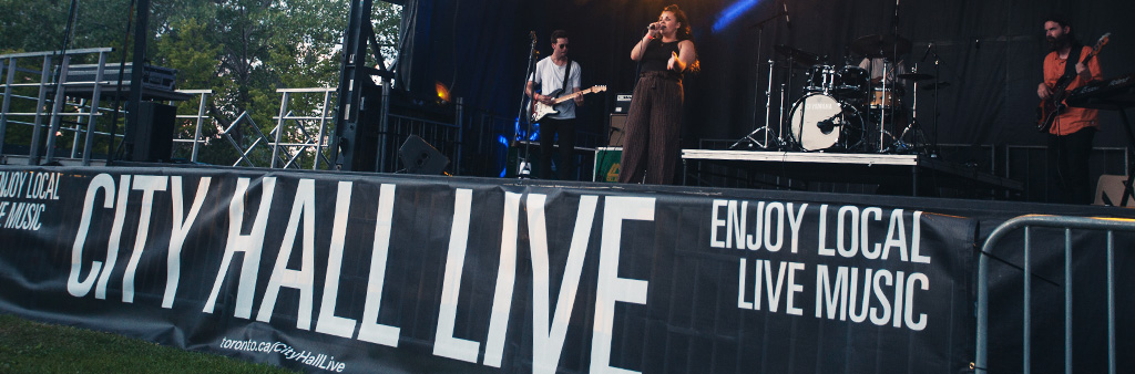 Band playing on outdoor stage at night, banner reads City Hall Live