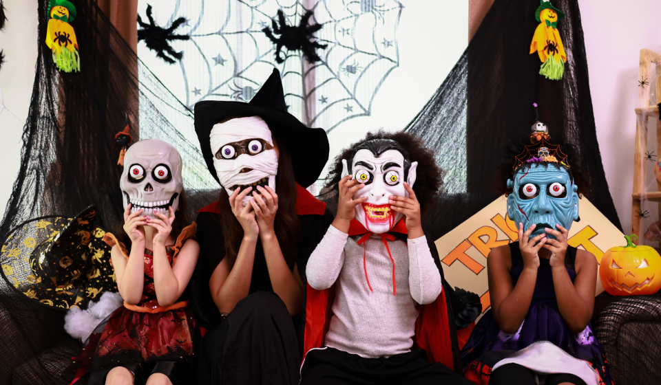 Three children and an adult cover their faces with monster masks while seated on a couch in room with Halloween decorations