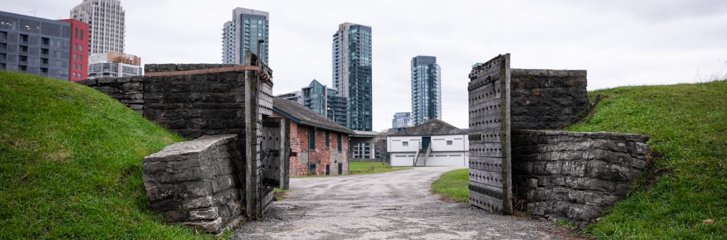 Path to historic fort with gate open showing block houses inside. Skyscrapers in the background.