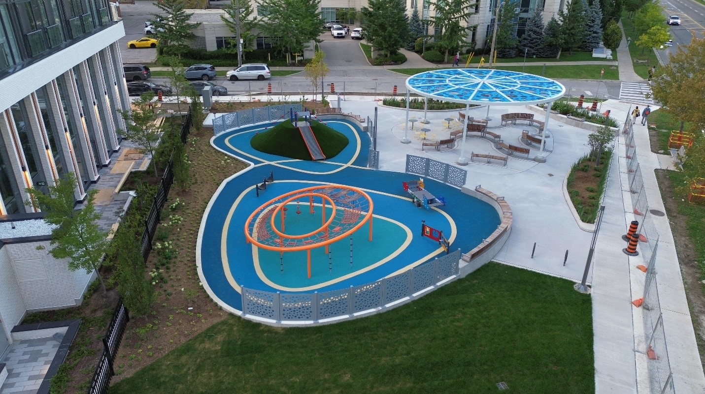 An image looking at the playground equipment including a circular and elevated climbing structure, and a hill slide located overtop rounded blue coloured rubber surfacing. A shade structure can be seen in the background with seating underneath. Fencing is placed around the park