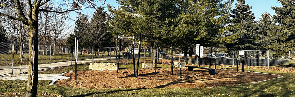 A fitness area with various pieces of equipment in black on top of mulch and surrounded by grass and a temporary fence.