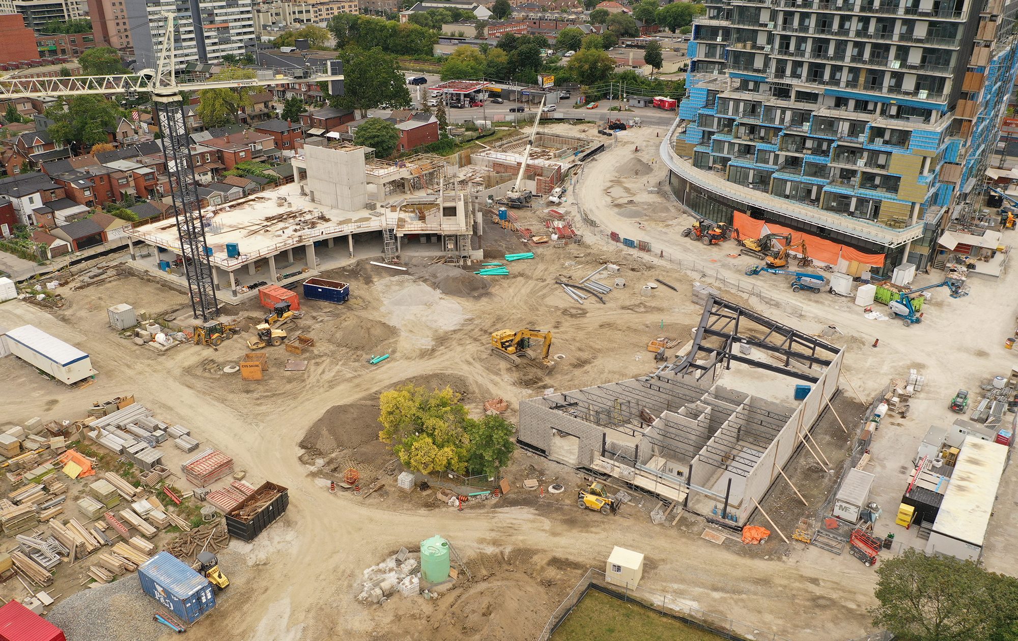 An aerial photograph of the construction site at Wallace Emersion Community Centre on September 10, 2023, which focuses on the community centre while under construction and the adjacent Fieldhouse. The community recreation structure is one-storey with vertical beams being installed to the future third floor. The Fieldhouse is at ground-level with concrete walls and steel structural beams. 