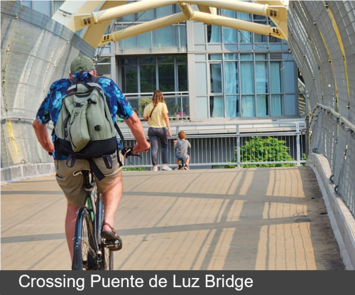 Person biking crosses the Puente de Luz Bridge. Text band across the bottom of the image reads “Crossing Puente de Luz Bridge”