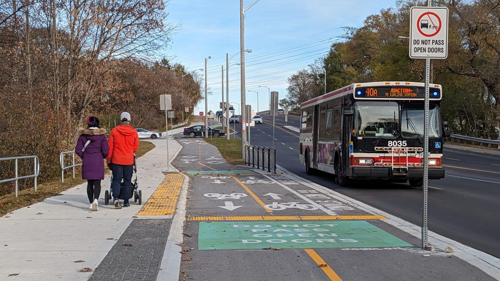 People walk on a sidewalk beside a two-way bikeway at the same level, with a TTC bus travelling in the motor vehicle travel lane to the right. 