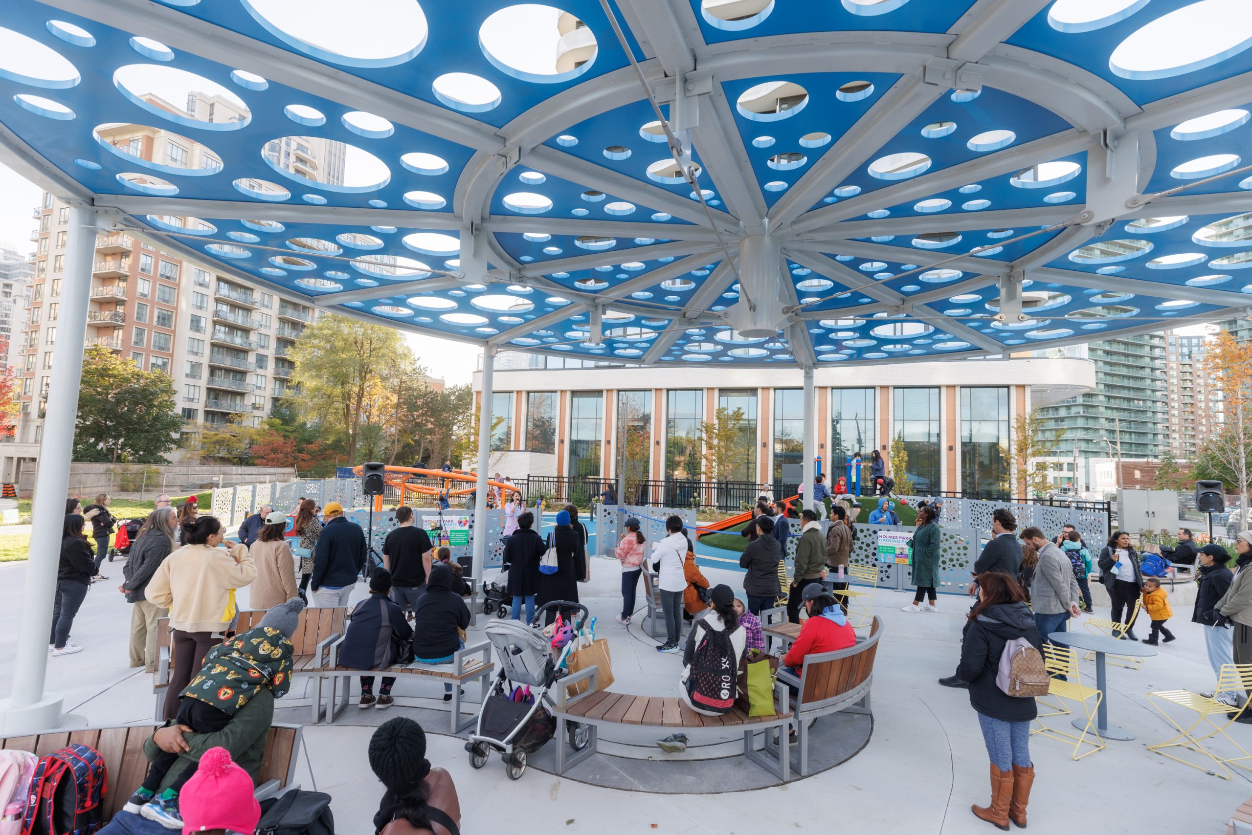 A view of the official park opening event during a keynote speech given by the local Councillor. A crowd of people are oriented towards the play space where the Councillor is standing.