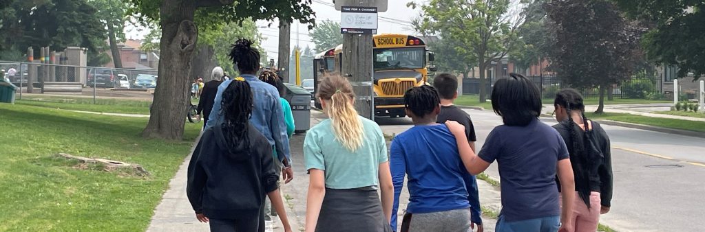 A large group of school children and at least one teacher are seen from behind as they walk close together along the sidewalk. In front of them, two school buses are parked in the curb lane. The grass and trees are lush and green, and the group of children are either wearing short sleeves or light layers.