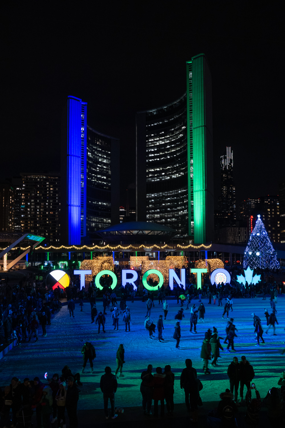 Outdoor skating rink with Toronto sign lit up, a giant Christmas tree and City Hall lit green and blue in the background.