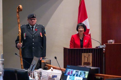 Presentation in Council for Indigenous Veterans Day on November 8, 2023. Mayor Chow is speaking at the podium, and behind her is Corporal Jason Nakogee of the Algonquin Regiment holding the Frances Pegahmagabow Eagle Staff