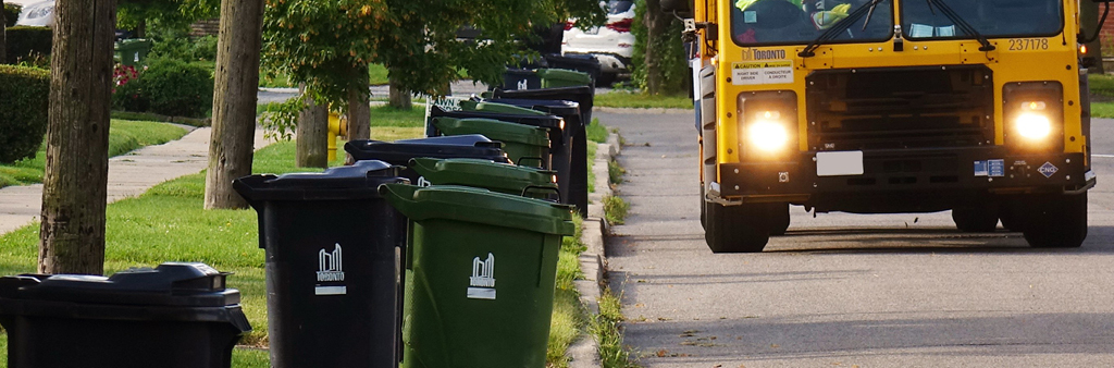 A City of Toronto collection truck drives down a residential street with collection bins at the curb.