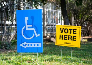 A blue voting sign with an accessible wheelchair logo on it and a yellow vote sign with the words Vote Here on a lawn.