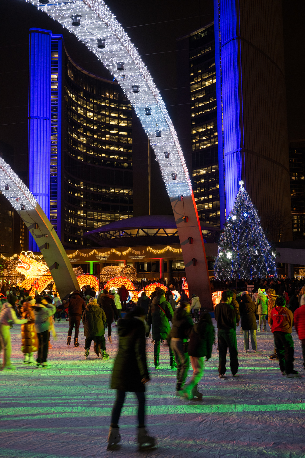 Lots of people skating on an outdoor skating a giant Christmas tree and City Hall lit blue in the background.
