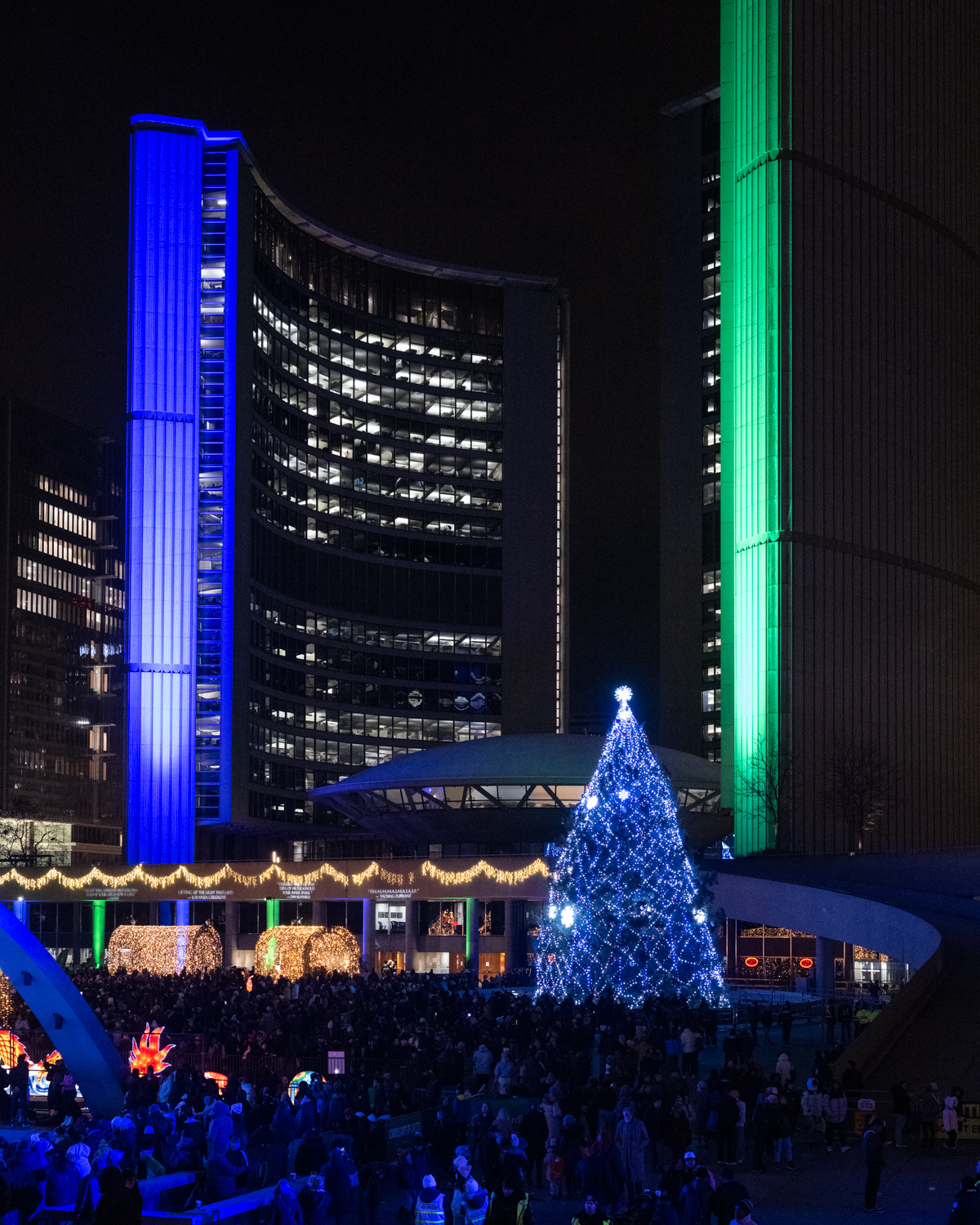 Toronto City Hall lit up blue and green at night with a giant glowing Christmas tree in front.