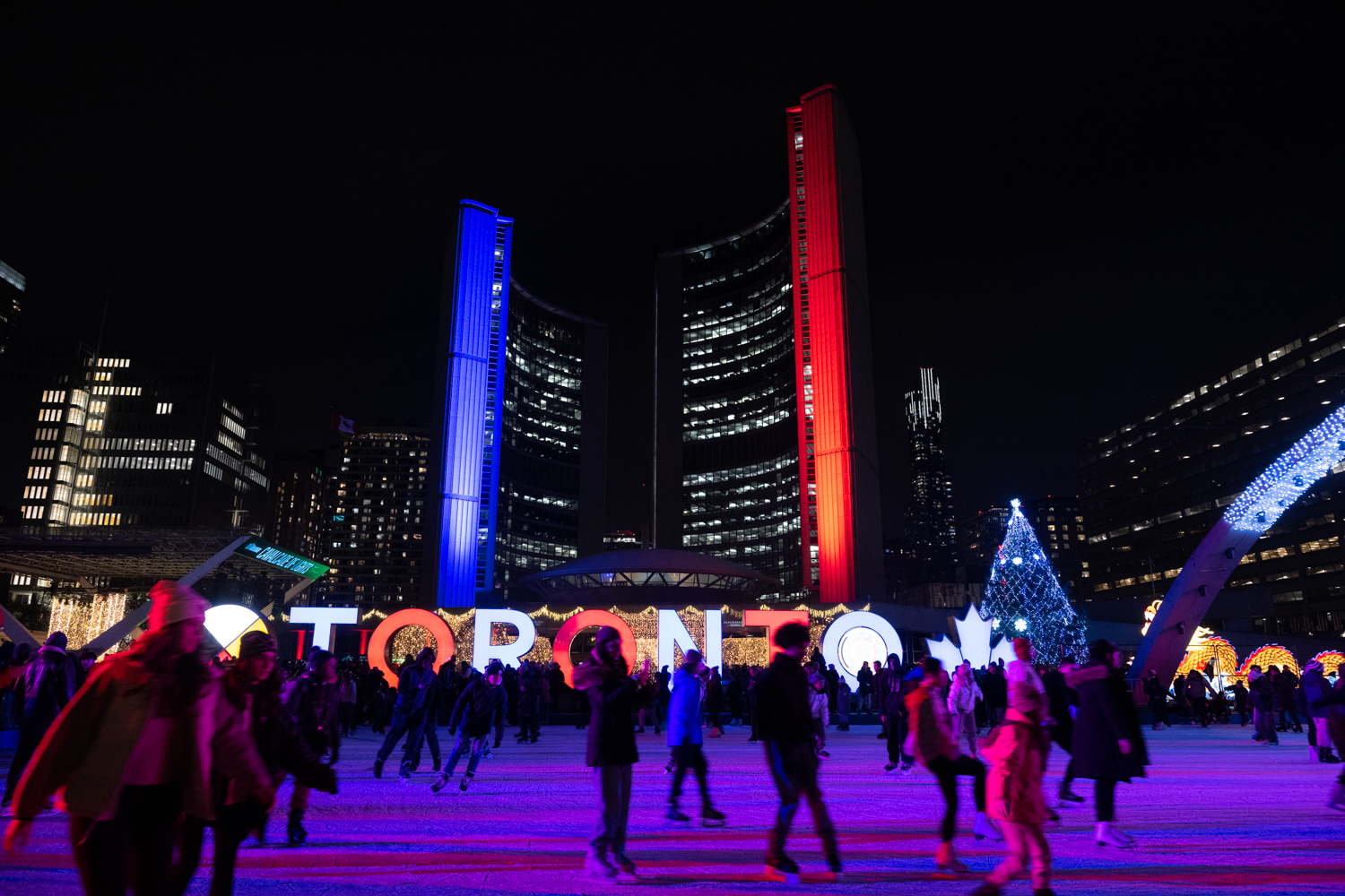 People skating outside in Nathan Phillips Square with Toronto sign lit up, a giant Christmas tree and City Hall lit red and blue in the background.