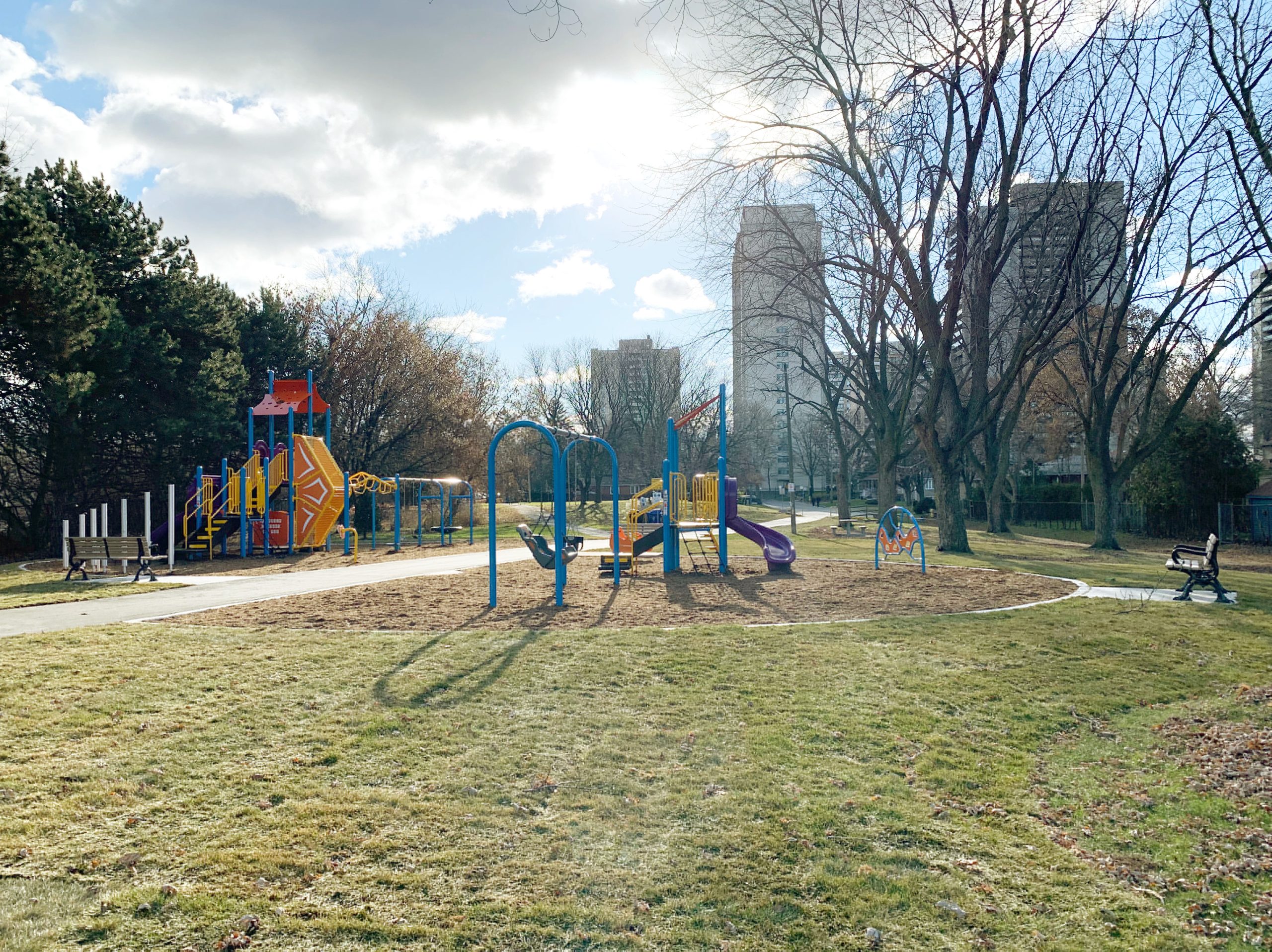 Benner Park Playground looking south. The playground is seperated into two areas by the central walkway; the senior play area is to the east of the walkway and the junior play area is to the west of the walkway. There is a mature tree in the foreground with more mature trees to the west and east behind both areas of the playground.