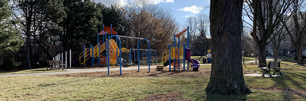 Benner Park Playground looking south-east. The playground is seperated into two areas by the central walkway; the senior play area is to the east of the walkway and the junior play area is to the west of the walkway. There is a mature tree in the foreground with more mature trees to the west and east behind both areas of the playground.