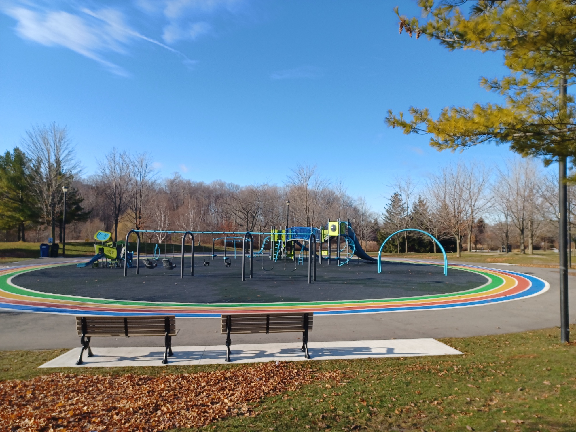 Milliken Park Playground after improvements which shows two benches in the foreground facing the new playground area which is circular in shape with equipment in blue and green. The playground area is surrounded by asphalt painted in rainbow colours.