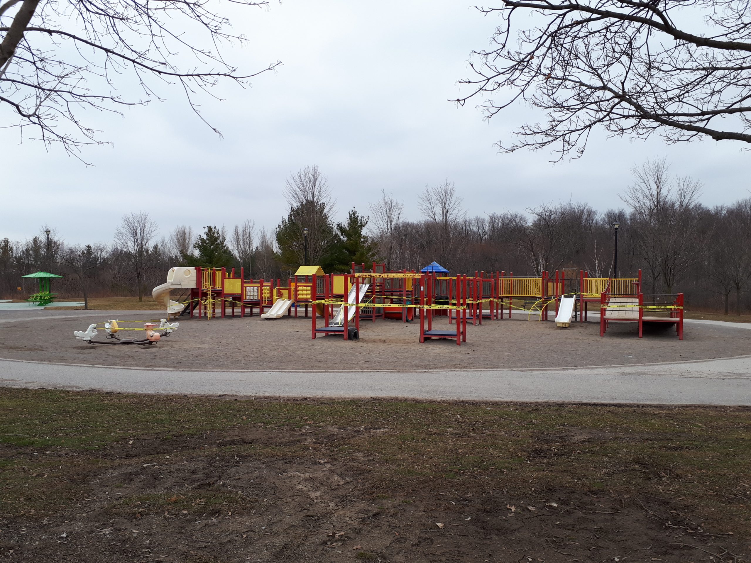 Banbury Park Playground which shows playground equipment in red and yellow on top of sand. The playground area is circular in shape and surrounded by a concrete pathway. The playground equipment is wrapped in caution tape as this photo was taken during the COVID-19 pandemic.