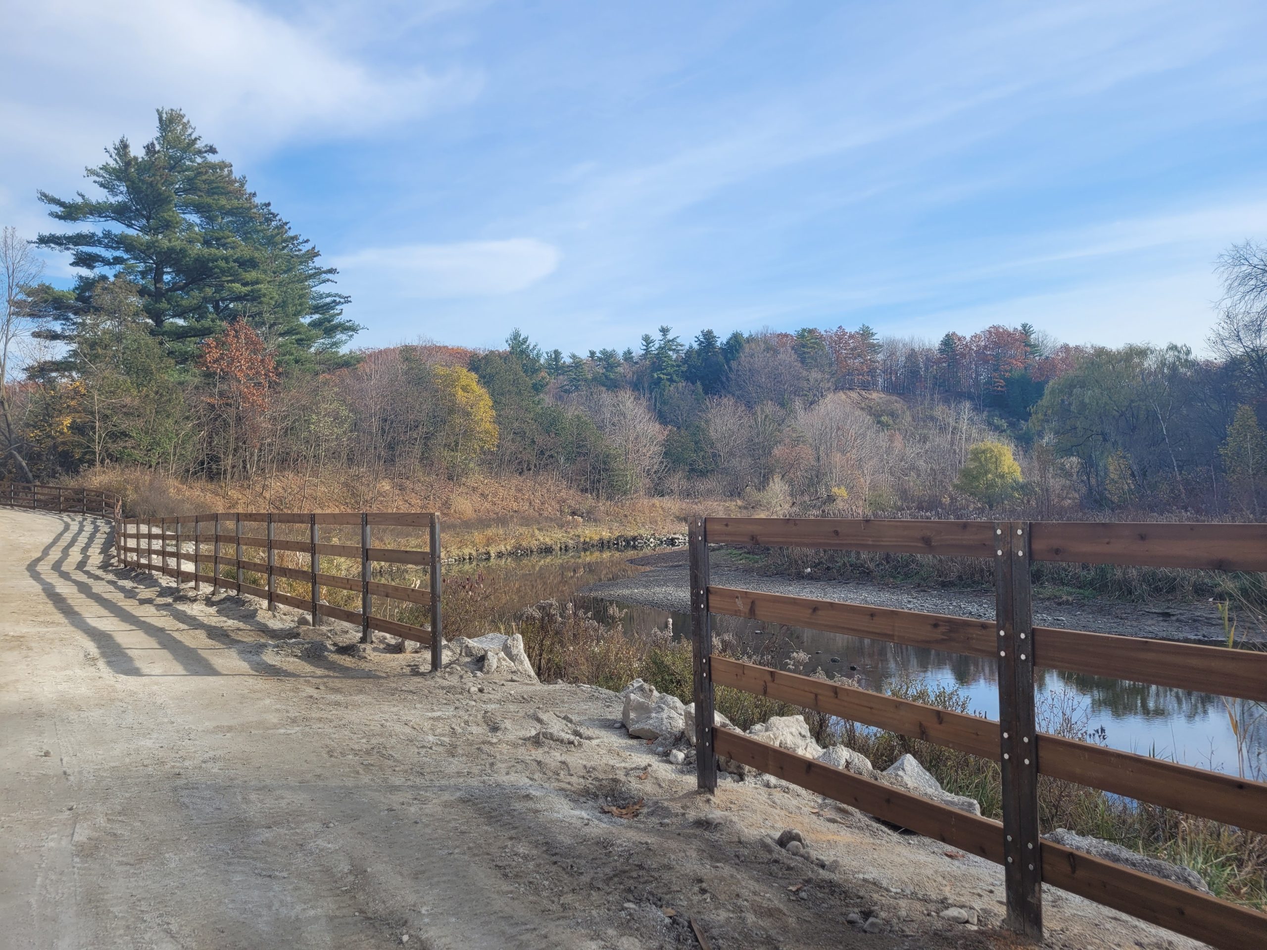 Photo of Highland Creek Trail during Phase 3A construction showing an unpaved trail path next to a pond divided by a wood fence under construction. Mature fall coloured trees are in the background. 