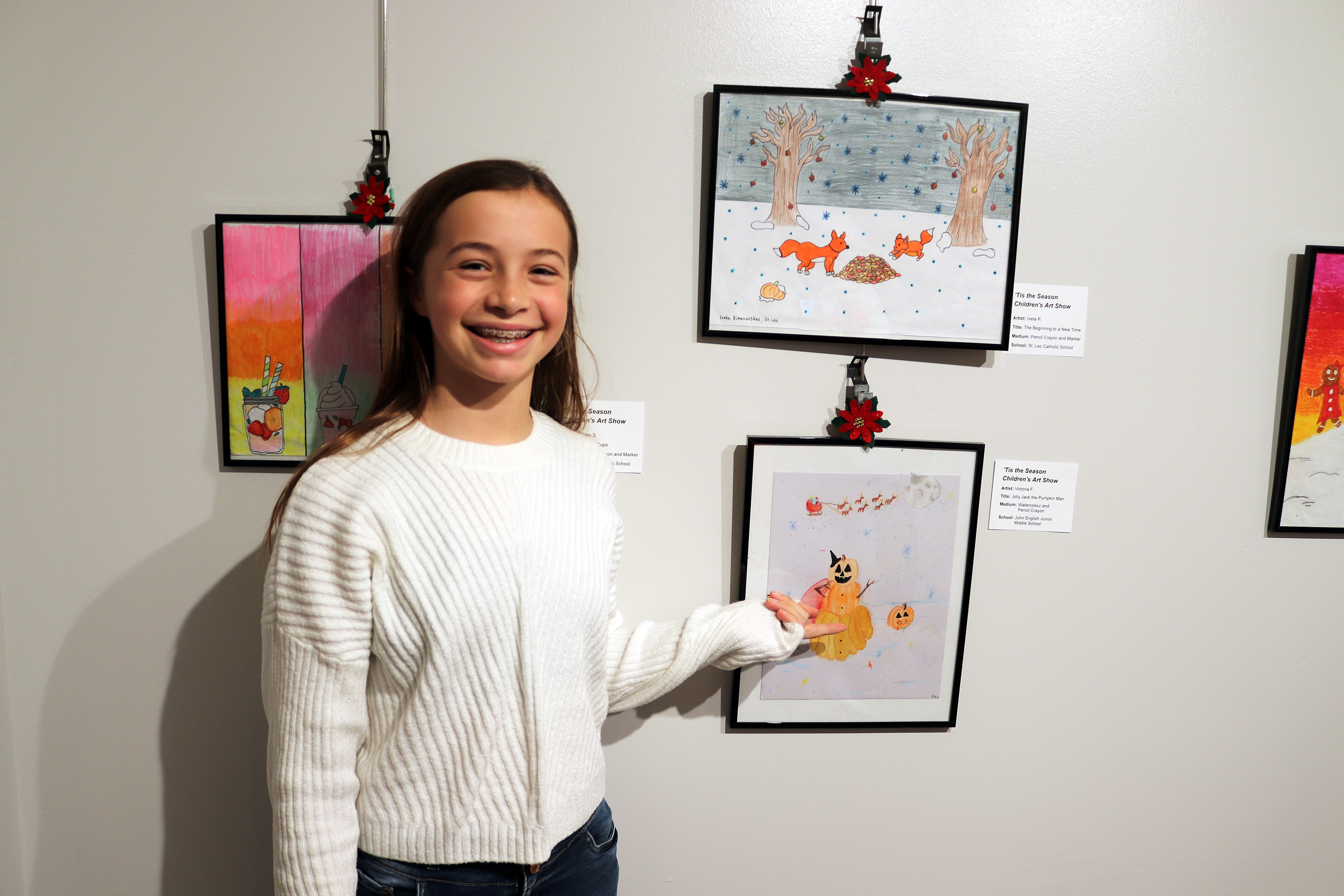 A student wearing a white sweater standing beside artwork on the walls and pointing to one of them
