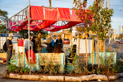 People gathered at strip mall patio with awning and plants.