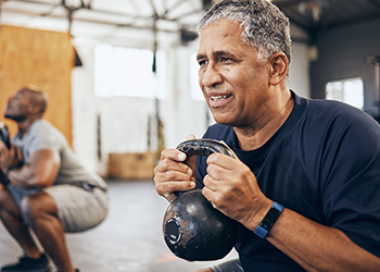 Close-up of an older adult holding a kettlebell at chest height with another person doing the same in the background.