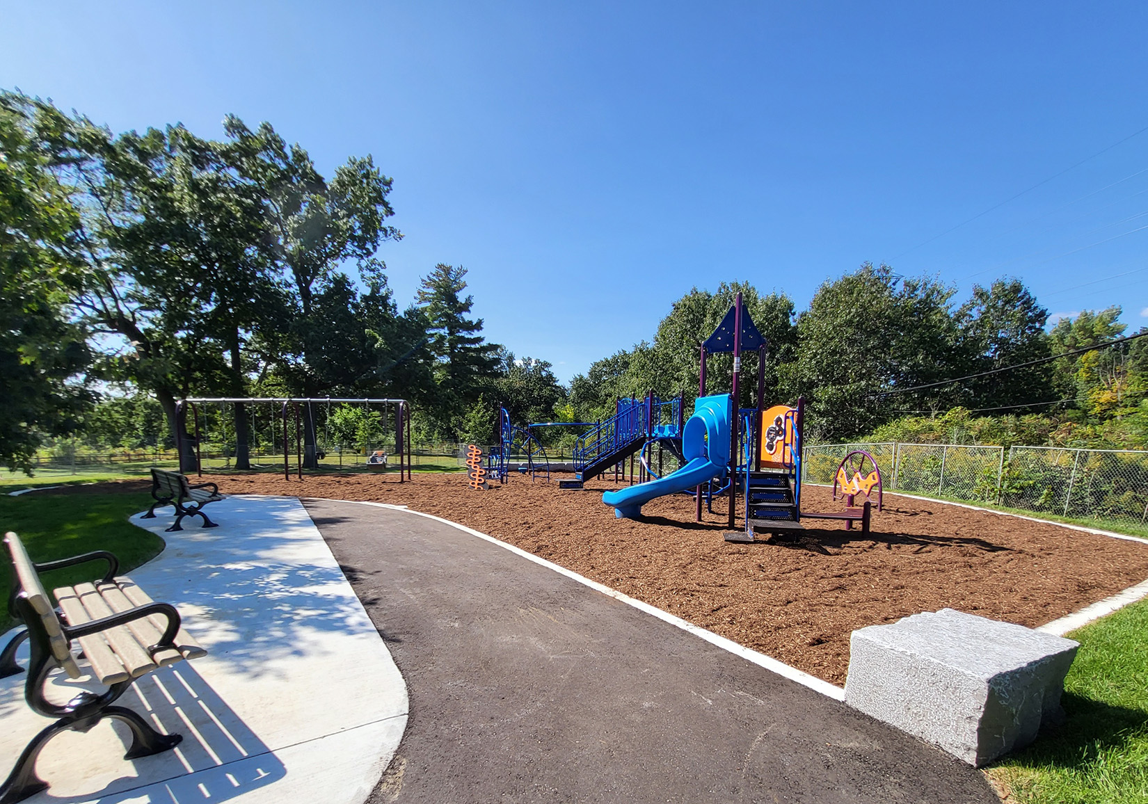 Photo of the new Lambton Park playground looking west. There is a seating area with two park benches, armourstone seating, and a junior play structure with a curved slide in the foreground, and a swing set, a stand-alone climber, and a senior play structure visible in the background.