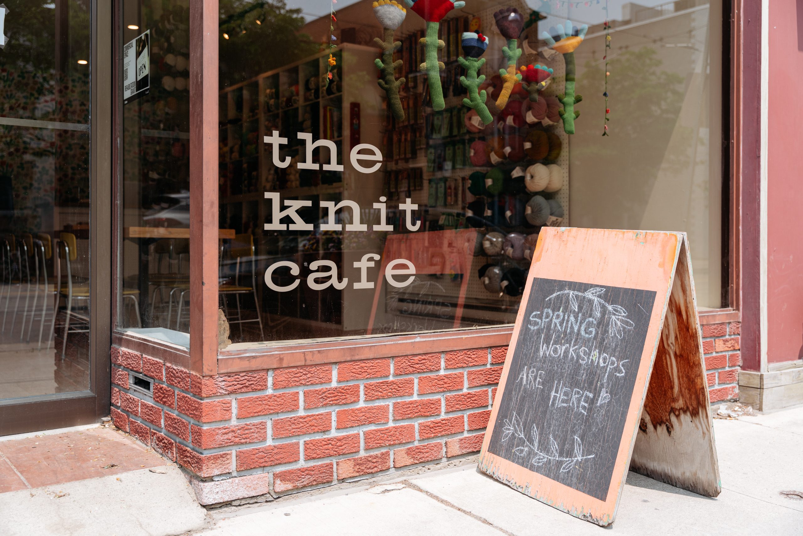 Exterior entrance of a shop named "The Knit Cafe" A-frame chalkboard sign reads "Spring Workshops Are Here."